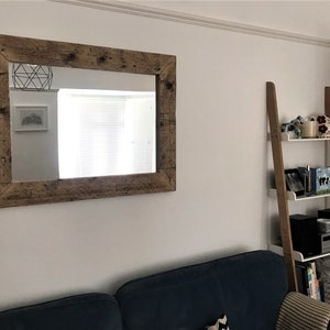 Op de afbeelding: Een grote rechthoekige spiegel met een rustieke houten lijst hangt aan een witte muur. De spiegel reflecteert een kamer met een raam en een wit plafond. Rechts in de afbeelding staat een witte ladderplank met drie planken. De planken zijn gevuld met verschillende items, waaronder een mok, een boek en een kleine luidspreker.