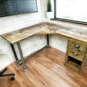 Puede incluir: Un escritorio de esquina hecho de madera recuperada con un marco de tubería de metal. El escritorio tiene dos cajones y un estante. El escritorio está en una habitación con paredes de ladrillo blanco y pisos de madera.