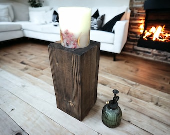 Op de afbeelding: Een donkerbruine houten kaarsenhouder met een witte kaars met gedroogde bloemen erin. De kaarsenhouder staat op een lichte houten vloer.