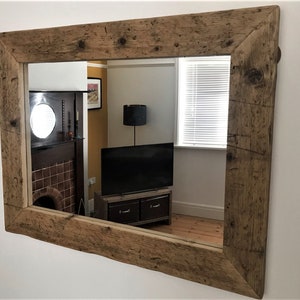 Op de afbeelding: Een grote rechthoekige spiegel met een rustieke houten lijst. De lijst is gemaakt van gerecycled hout met een natuurlijke bruine afwerking. De spiegel reflecteert een kamer met een open haard, een televisie en een raam.