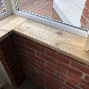 May include: A close-up of a wooden windowsill with a light brown finish. The windowsill is made of reclaimed wood and has a rustic look. The windowsill is installed on a brick wall.