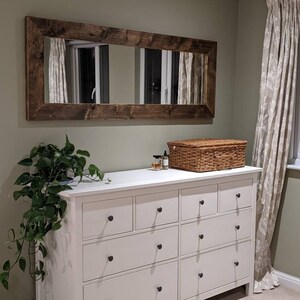 Op de afbeelding: Een witte commode met acht laden en zwarte knoppen. De commode staat voor een groene muur met een grote rechthoekige spiegel met een donkerhouten lijst. Een rieten mand en een paar flessen staan op de commode.