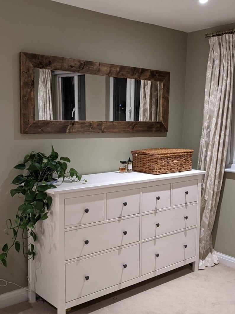 Op de afbeelding: Een witte commode met acht laden en zwarte knoppen. De commode staat voor een groene muur met een grote rechthoekige spiegel met een donkerhouten lijst. Een rieten mand en een paar flessen staan op de commode.