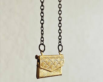 May include: A gold-toned metal pendant shaped like a small envelope or clutch purse, featuring an embossed repeating scalloped pattern on the front flap. The pendant hangs from a dark, oxidized metal chain with circular links on both sides.