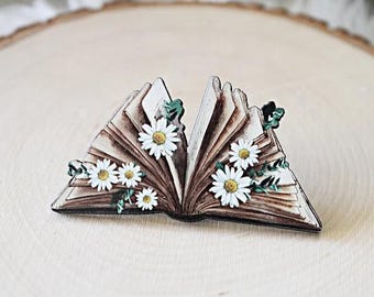 May include: A decorative pin shaped like an open book with aged, brown-toned pages. The book is adorned with several white daisy flowers featuring yellow centers and small green leaves tucked between the pages, creating a detailed floral design.