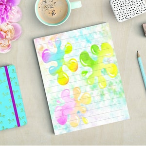 May include: A lined notepad with colorful abstract paint splatters in shades of pink, yellow, green, and blue. The notepad is on a light wooden surface, alongside pencils, a turquoise journal, and a cup of coffee.