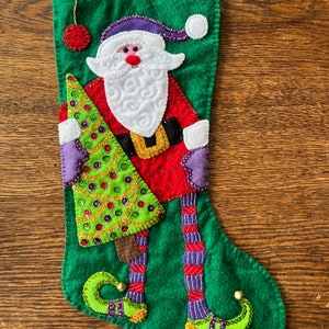 Puede incluir: Calcetín de Navidad de fieltro verde con un diseño de Papá Noel. Papá Noel sostiene un árbol de Navidad verde con lentejuelas doradas y verdes. Papá Noel lleva un traje rojo y blanco con un gorro morado y botas moradas.