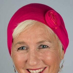 May include: A woman with short, white hair is smiling. She is wearing a bright pink hat with a pink flower on the side. The hat is made of a soft, stretchy fabric.