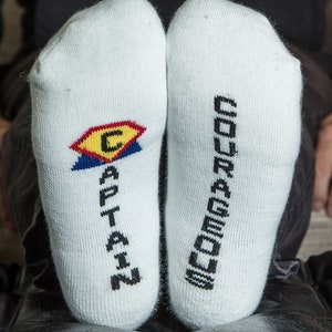 May include: A pair of white socks with black text. One sock says "CAPTAIN" with a red, yellow, and blue logo. The other sock says "COURAGEOUS".