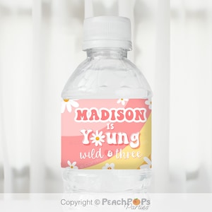 May include: Water bottle label with a pink and yellow background featuring white daisies. The label says "Madison is Young wild & three" in a retro font.