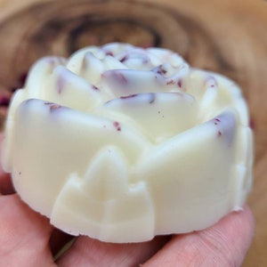 May include: A white rose-shaped soap with pink and red flecks throughout. The soap is in the shape of a rose with a detailed design.