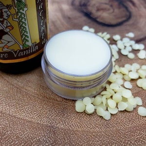 May include: A small clear jar filled with a white lip balm, next to a bottle of pure vanilla extract and a pile of small, round, yellow wax beads.
