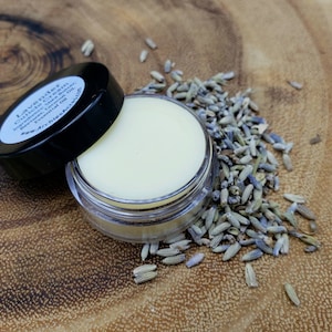 May include: A small clear jar with a white lid containing a white balm. The jar is open and the balm is visible. Dried lavender flowers are scattered around the jar on a wooden surface. The jar has a white label that reads "Lavender Citrus Shea Butter Infused Coconut Oil Essential Oil" and "www.archiessshop.com".