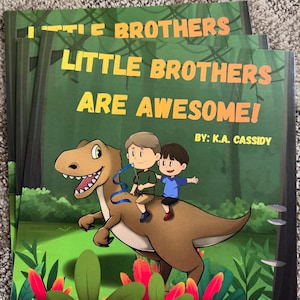 Pode incluir: Um livro ilustrado infantil intitulado Little Brothers Are Awesome! de K.A. Cassidy. A capa apresenta uma ilustração de desenho animado de dois meninos montados em um tiranossauro marrom em uma floresta com plantas tropicais.