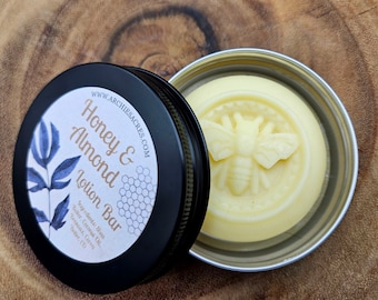 Może przedstawiać: Okrągłe puszeczka z czarnym wieczkiem. Wieczko ma białą etykietę z tekstem "WWW.ARCHIESACRES.COM Honey & Almond Lotion Bar" i grafiką niebieskiego liścia. Pudełko jest otwarte, a wewnątrz widoczny jest okrągły, biały batonik do ciała z wzorem pszczoły.