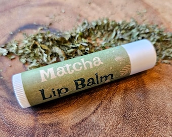Könnte beinhalten: Eine weiße Tube Lippenbalsam mit einem grünen Etikett, auf dem "Matcha Lip Balm" steht. Das Etikett hat ein grünes und weißes Design mit einem Blattmotiv. Der Lippenbalsam liegt auf einer Holzoberfläche mit verstreuten grünen getrockneten Blättern.