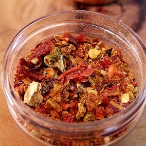 May include: A clear glass jar filled with a mix of dried red chili flakes, green herbs, and yellow spices. The jar is sitting on a wooden surface, and the contents are in sharp focus.