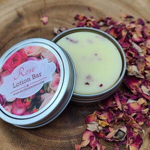 May include: A small tin container with a white lotion bar inside. The tin has a label that says "Rose Lotion Bar" and "www.ArchiesAcres.com". The lotion bar is surrounded by dried rose petals.