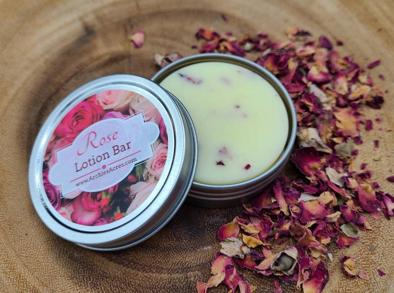 May include: A small tin container with a white lotion bar inside. The tin has a label that says "Rose Lotion Bar" and "www.ArchiesAcres.com". The lotion bar is surrounded by dried rose petals.