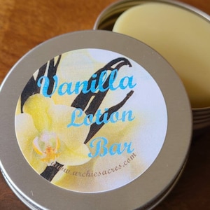 May include: A silver tin with a white label that says "Vanilla Lotion Bar" with a picture of a white orchid and vanilla beans. The tin contains a round, white lotion bar.