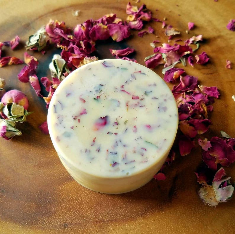 May include: A round, white soap bar with dried rose petals embedded in it. The soap is sitting on a wooden surface.