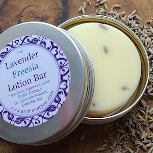 May include: A 2 ounce tin of lavender freesia lotion bar. The tin is silver with a purple and white label that reads "Lavender Freesia Lotion Bar" and lists the ingredients: beeswax, shea butter, coconut oil, lavender flowers, and essential oils. The lotion bar is white and has a few dried lavender flowers on top.