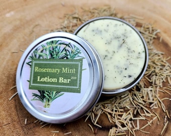 May include: A silver tin with a white label that reads "Rosemary Mint Lotion Bar 2 oz". The label features a green illustration of rosemary sprigs. The tin is open and a white lotion bar with green flecks is visible inside.