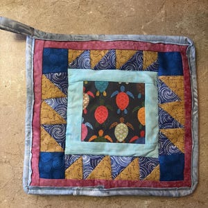 May include: Square quilted potholder with a loop for hanging. The center features a dark background with colorful turtles. The design includes blue, gold, and pink patterned fabric, creating a vibrant and functional kitchen accessory.