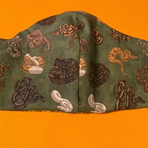 May include: A green fabric face mask with a repeating pattern of brown and white snakes. The mask has elastic ear loops.