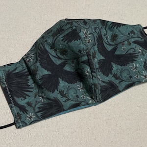 May include: A dark green face mask with a black raven pattern. The mask has a black elastic band.