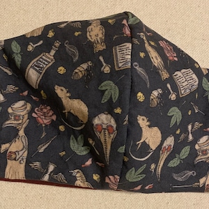 May include: A dark gray face mask with a repeating pattern of illustrations including rats, roses, and plague doctor figures. The mask has black straps for securing it to the face.