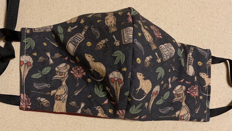 May include: A dark gray face mask with a repeating pattern of illustrations including rats, roses, and plague doctor figures. The mask has black straps for securing it to the face.