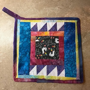 May include: A square quilted potholder featuring a central black fabric panel with white unicorn illustrations, rainbow accents, and the text "I'm a Unicorn Bitch Please". The border consists of purple, orange, blue, and yellow patterned fabric strips.