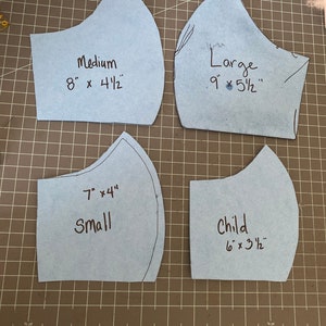 May include: Four light blue face mask patterns in various sizes: Medium (8" x 4.5"), Large (9" x 5.5"), Small (7" x 4"), and Child (6" x 3.5"). Each pattern is labeled with its size and dimensions in inches. The patterns are on a gray cutting mat.