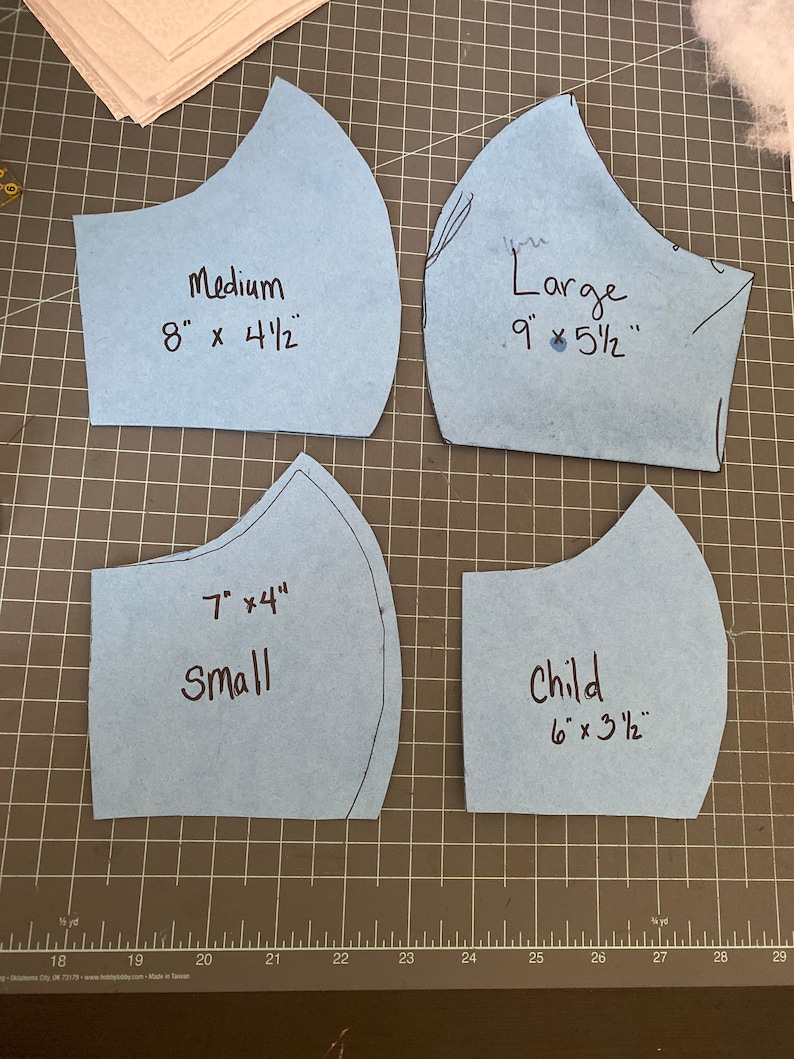 May include: Four light blue face mask patterns in various sizes: Medium (8" x 4.5"), Large (9" x 5.5"), Small (7" x 4"), and Child (6" x 3.5"). Each pattern is labeled with its size and dimensions in inches. The patterns are on a gray cutting mat.