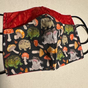May include: A handmade face mask featuring a whimsical pattern of frogs and mushrooms on a black background. The mask has a red patterned lining and black elastic ear loops. The design includes orange, green, and brown elements.