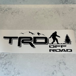 May include: Black and white decal with the text "TRD OFF ROAD" and a silhouette of a mountain range, a Bigfoot, and a pine tree.