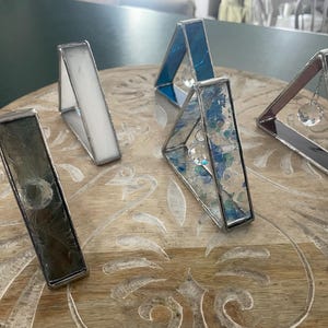 May include: A collection of handmade stained glass triangle-shaped candle holders in various colors. The candle holders are made with clear, white, blue, and maroon glass, and some feature a hanging crystal. They are displayed on a wooden surface.