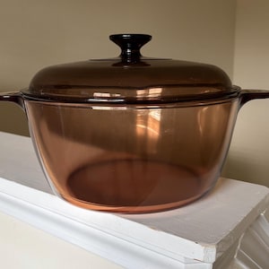 May include: A round, amber-colored glass casserole dish with a matching translucent lid. The pot features two integrated side handles and a central knob on the lid. The glass is smooth and transparent, allowing light to pass through the warm brown tint.