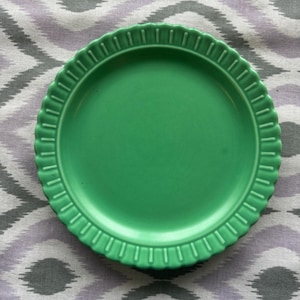 May include: A solid green ceramic plate with a scalloped edge. The plate is round and has a slightly raised rim. The plate is sitting on a patterned fabric with gray, white, and purple chevron stripes.
