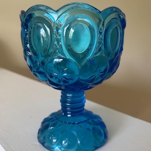 May include: A vintage, turquoise-colored glass pedestal bowl. The bowl features a scalloped rim and is adorned with star and oval patterns. The base is round with a starburst design, and the stem is ribbed.