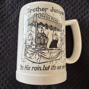 May include: White ceramic beer stein with a handle, featuring a black and white cartoon illustration of a parish picnic under a canopy. The text "Brother Juniper" and "Parish Picnic" are visible, along with the quote "It's His rain, but it's our picnic."