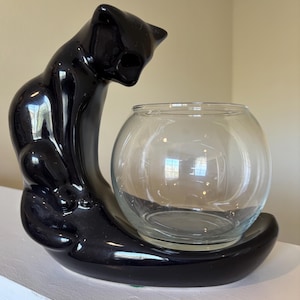 May include: A black ceramic cat-shaped candle holder with a glossy finish. The cat is arched over a clear glass bowl, creating a unique decorative piece. The base is a matching black color, providing a stable foundation for the bowl.