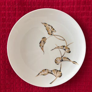 May include: White ceramic plate with a gold and black leaf design. The leaves are stylized and arranged in a graceful pattern across the plate's surface. The plate has a simple, elegant design.