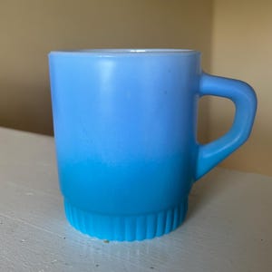 May include: A light blue mug featuring a smooth gradient that transitions from a pale sky blue at the top to a vibrant, saturated cyan at the base. The mug has a sturdy handle and a distinctively ribbed, vertical texture around the bottom edge.