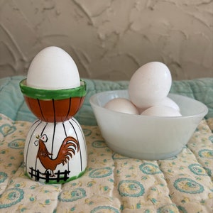 May include: A white ceramic egg cup with a rooster design. The egg cup has a green and orange band around the top and a white base with a rooster illustration. The rooster is brown and white with a red comb and wattle. The egg cup is sitting on a white and green floral patterned fabric. There are four white eggs in a white bowl behind the egg cup.