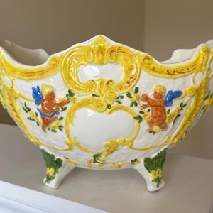 May include: A white ceramic bowl with a scalloped rim, featuring raised yellow scrollwork, green leaf accents, and two orange cherub figures with blue wings. The bowl is supported by three ornate feet decorated with green and yellow floral details.