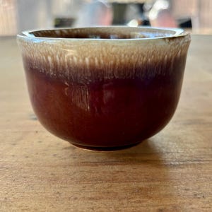 May include: A brown and white glazed ceramic cup with a rounded shape. The cup has a brown glaze with a white rim.