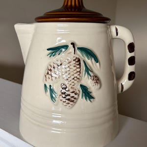 May include: A cream-colored ceramic pitcher with a brown lid, featuring a raised relief design of brown pinecones and green pine needles on the front. The handle is decorated with three brown spots, and the body has horizontal embossed lines.