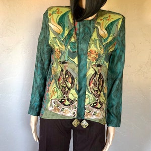 Puede incluir: Una chaqueta verde azulada con estampado floral con jarrones y flores. La chaqueta tiene un diseño largo y fluido y un escote sin cuello. La chaqueta está adornada con un collar largo verde oscuro con dos colgantes dorados.